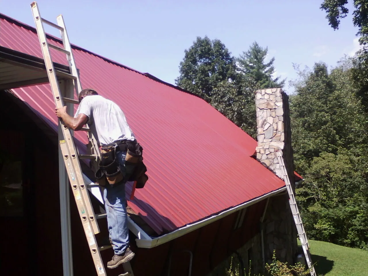 Roofer on a ladder beside a red metal roof and stone chimney — Soffit & Fascia Repair by Roofing Brothers in Dairy
